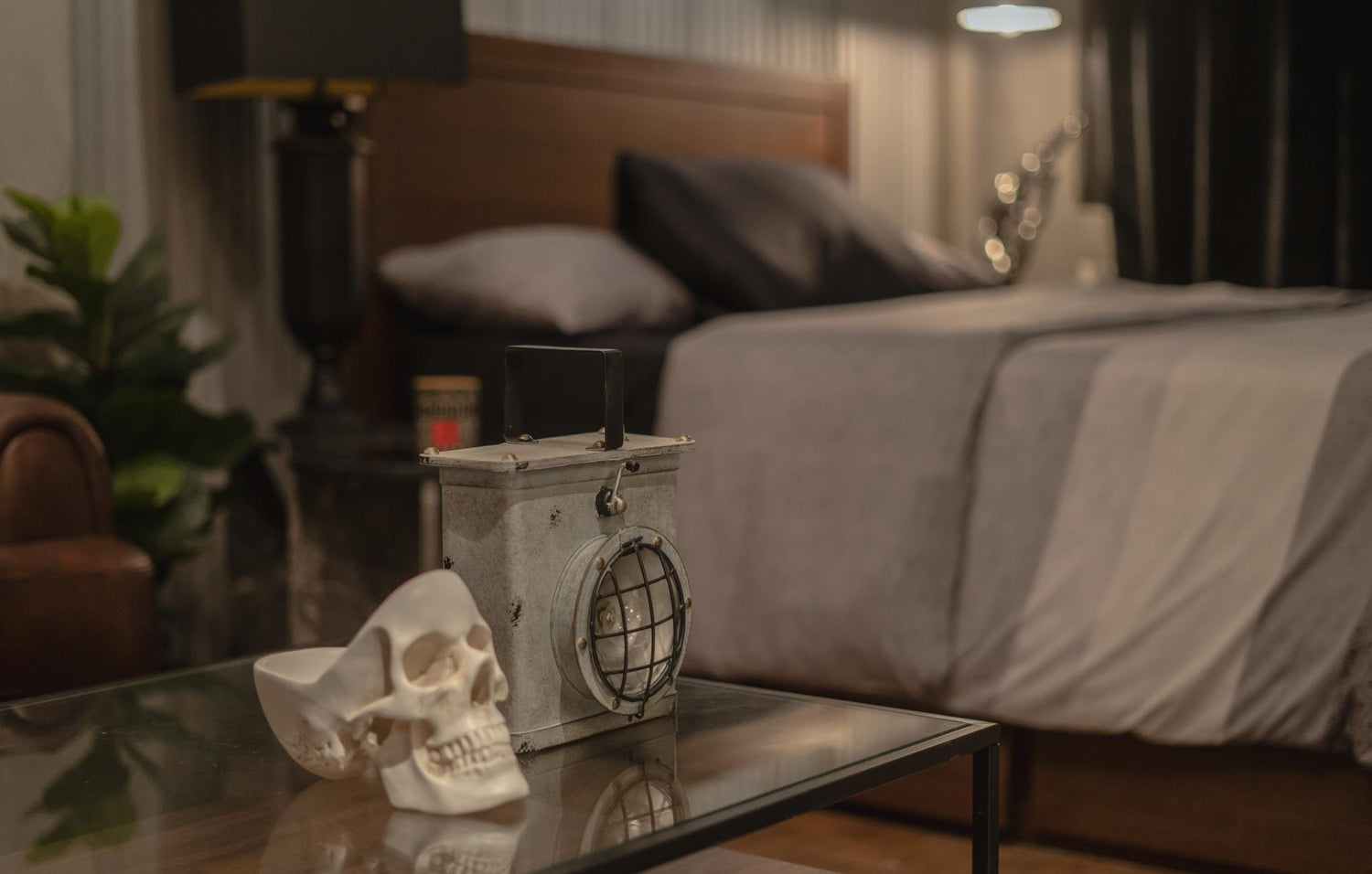 A human skull replica and an old lantern on a glass table in a cozy bedroom setting.