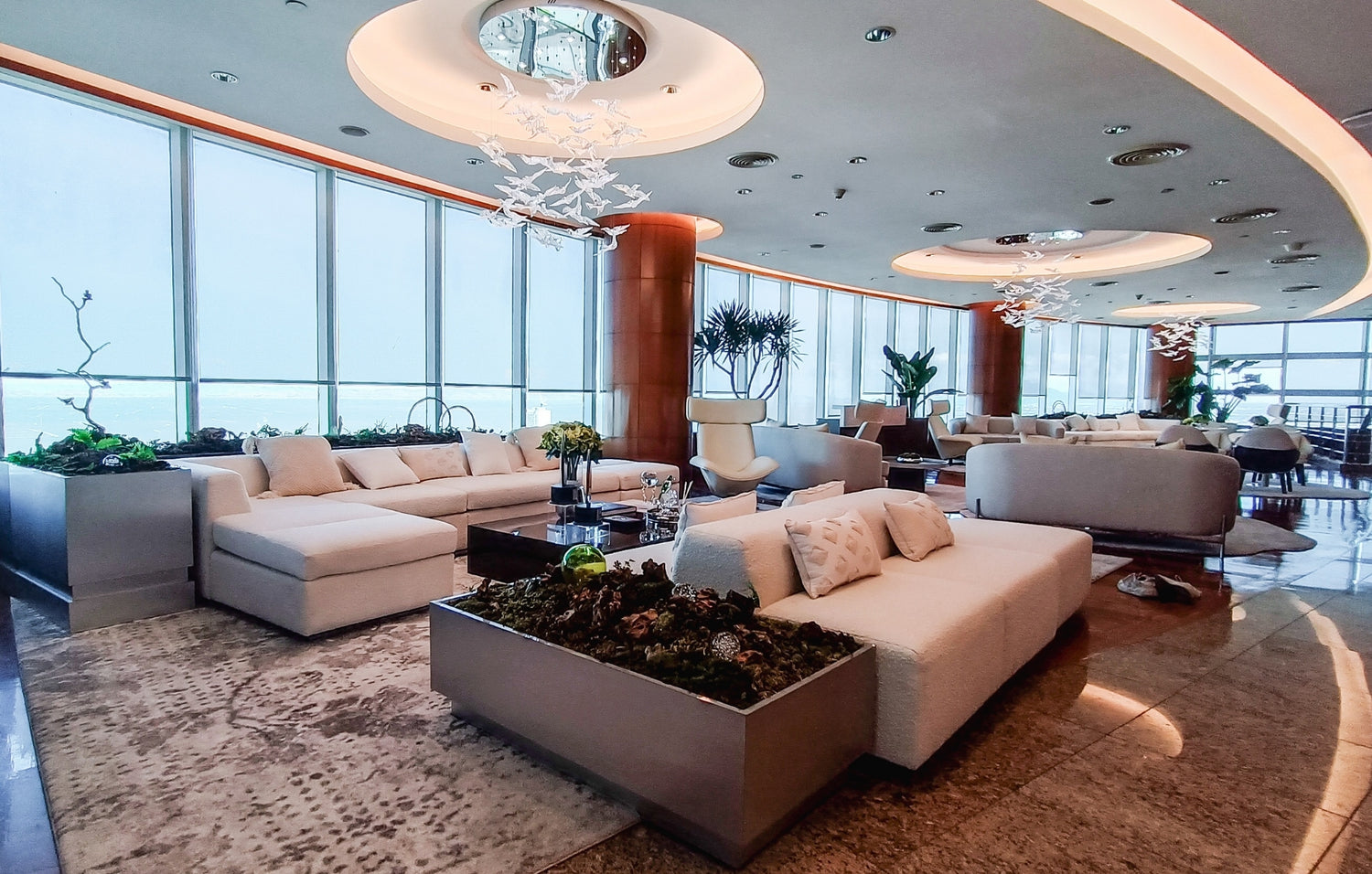 Modern lounge with white sofas, floor-to-ceiling windows, and artistic ceiling lights.