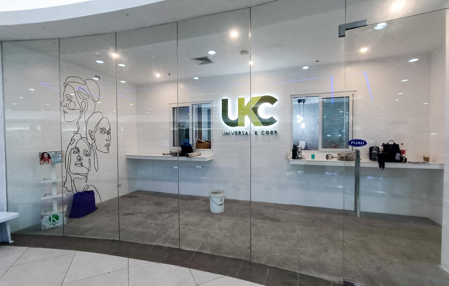 Glass-fronted office space with the logo ‘UKC Universal K Corp’ illuminated on the back wall. The interior includes minimalistic counters and a clean white design. Abstract line art of human faces is displayed on the left glass panel, with a gray tiled floor and scattered items visible inside.