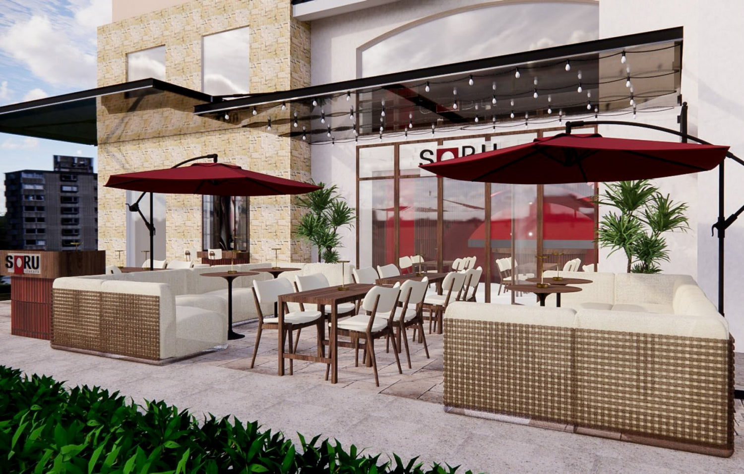 Outdoor patio area with comfortable wicker sofas, white cushions, wooden dining tables, and chairs. Red umbrellas provide shade, and string lights hang overhead. The background features a modern building with large glass doors labeled 'SORU,' stone accents, and potted plants.