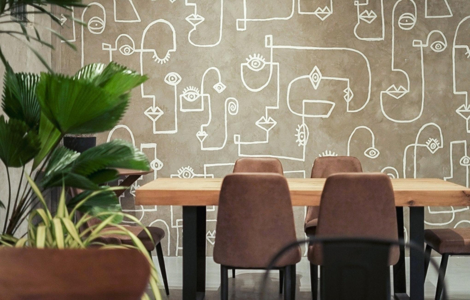 A dining area with a wooden table, brown chairs, green plants, and a wall with abstract line drawings.