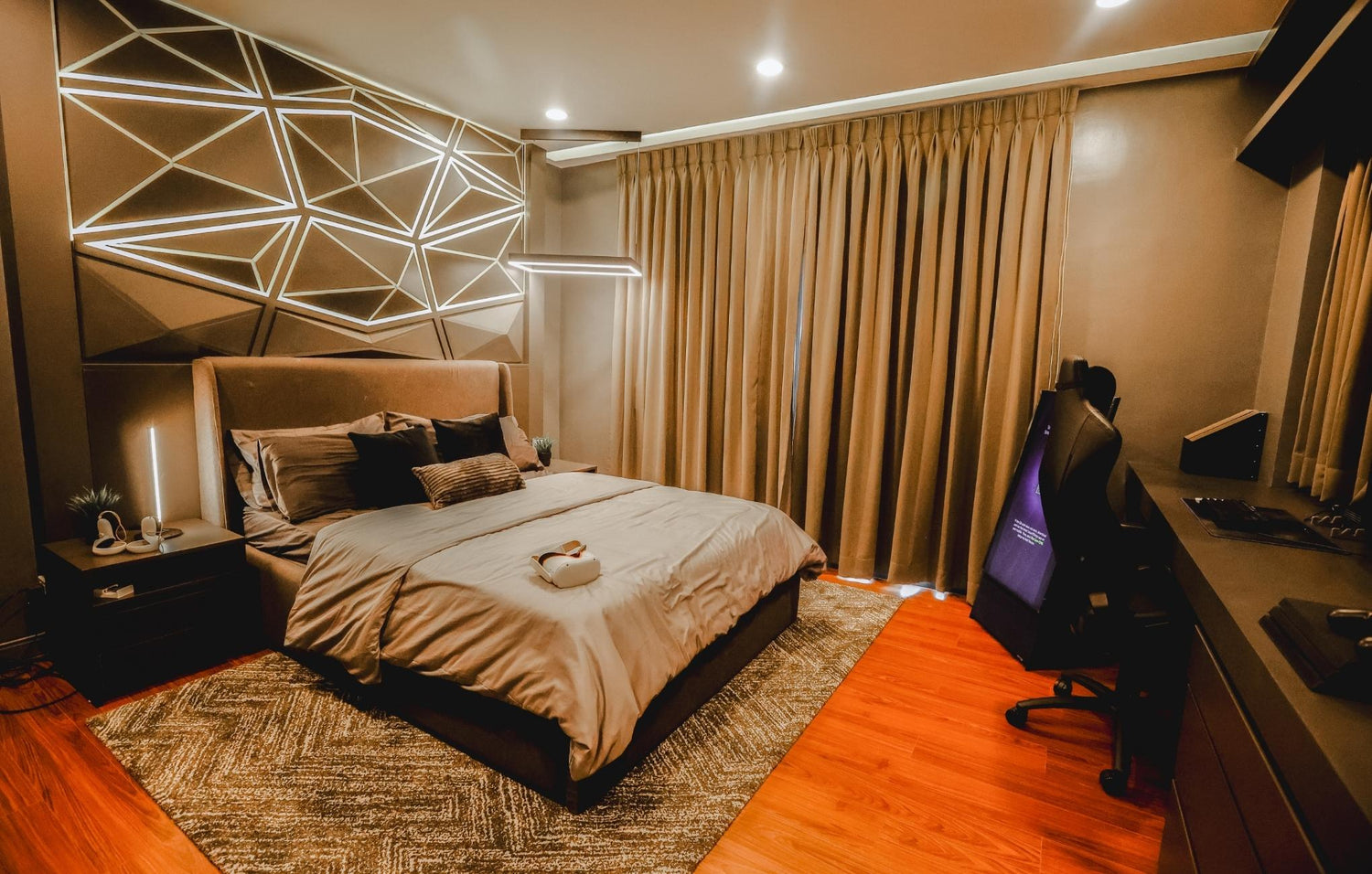 Modern bedroom with geometric accent lighting on the wall, a cozy upholstered bed, warm wood flooring, and a gaming setup in the corner."