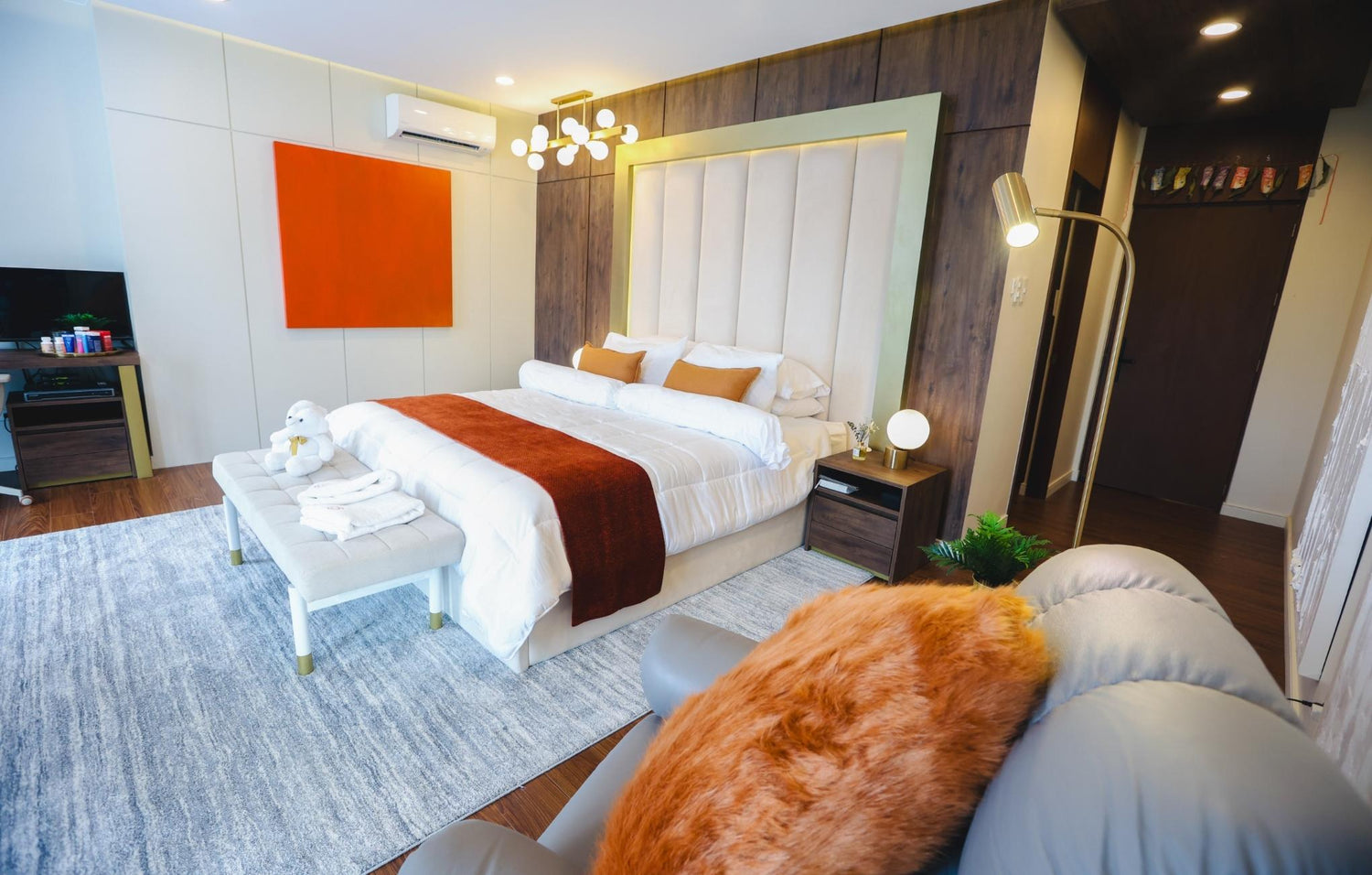 Luxurious bedroom featuring a tufted headboard, bold orange accents, a cozy throw, modern lighting fixtures, and a comfortable seating area with warm decor elements.