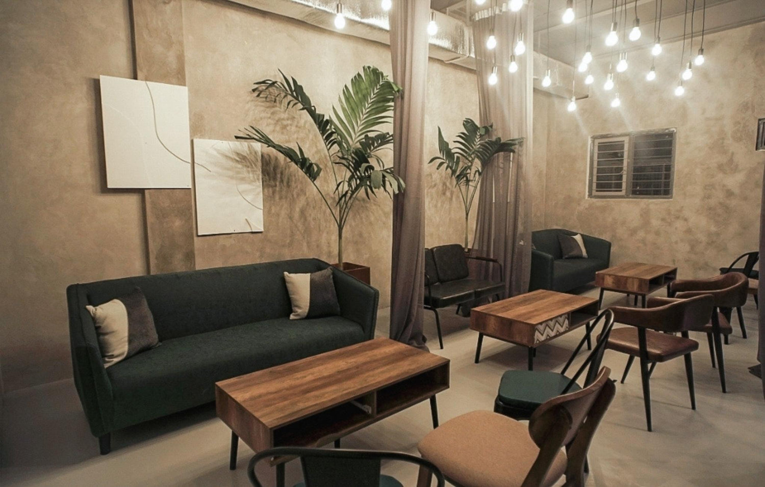 A cozy room with a dark green sofa, wooden tables, chairs, hanging lights, and wall art.