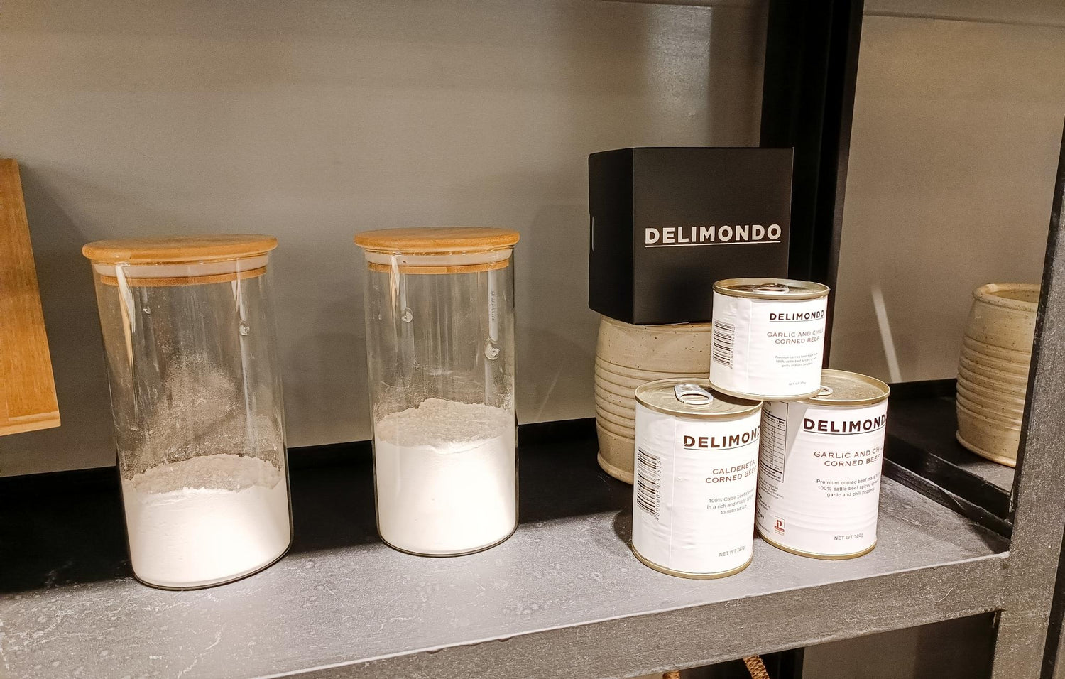 A kitchen shelf with two glass jars of flour and four cans of Delimondo brand corned beef.