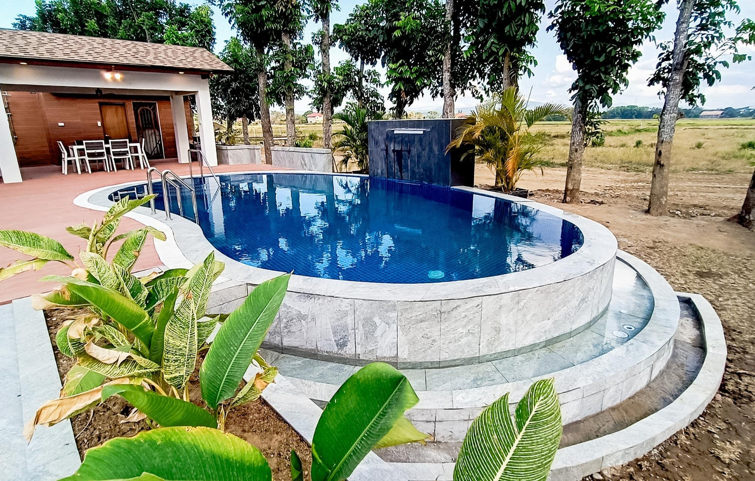 Swimming pool with curved edges near a wooden house, surrounded by greenery under a clear sky.