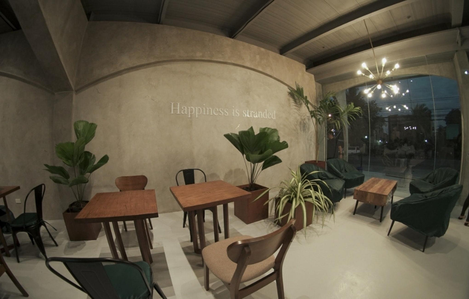 Interior of a stylish café with wooden tables, chairs, plants, and the phrase "Happiness is stranded" on the wall.