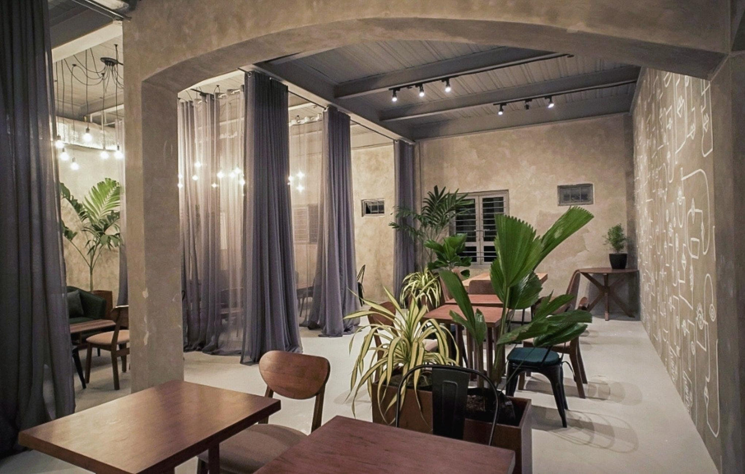 Elegant cafe interior with plants, wooden tables, draped curtains, and decorative lighting.