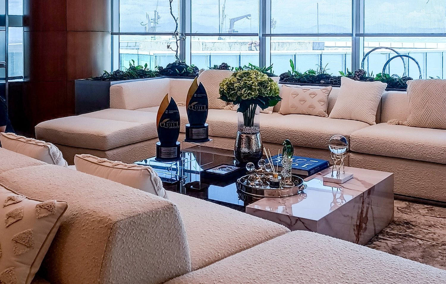 Elegant living room with beige sofas, marble table, decorative trophies, and a waterfront view.