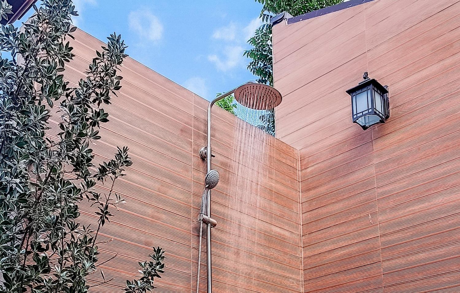 Outdoor shower with water flowing against a wooden wall, flanked by greenery and a wall lamp.
