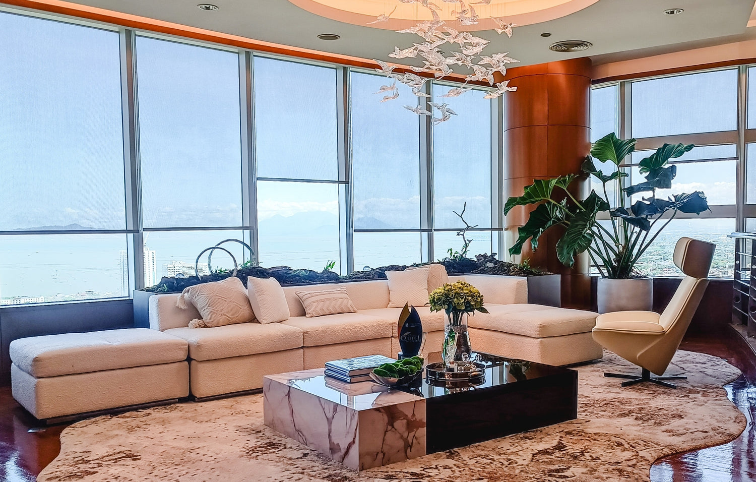 Modern living room with large windows overlooking the sea, beige sofa, marble table, and indoor plants.