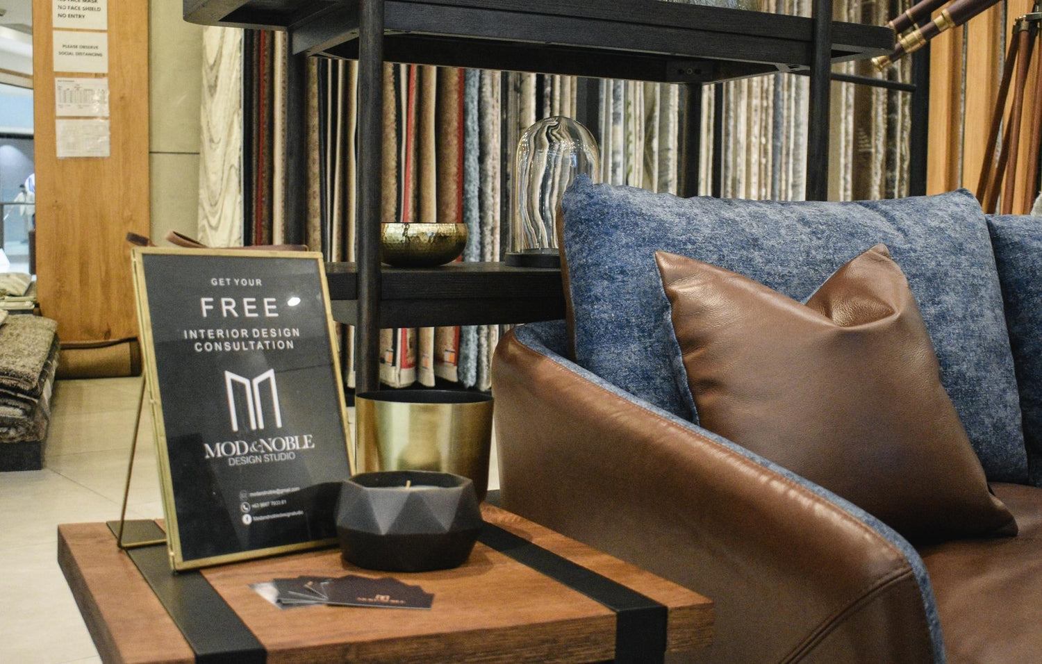 Interior design studio advertisement on easel, with leather chair, decorative pillows, and shelves with fabric samples.