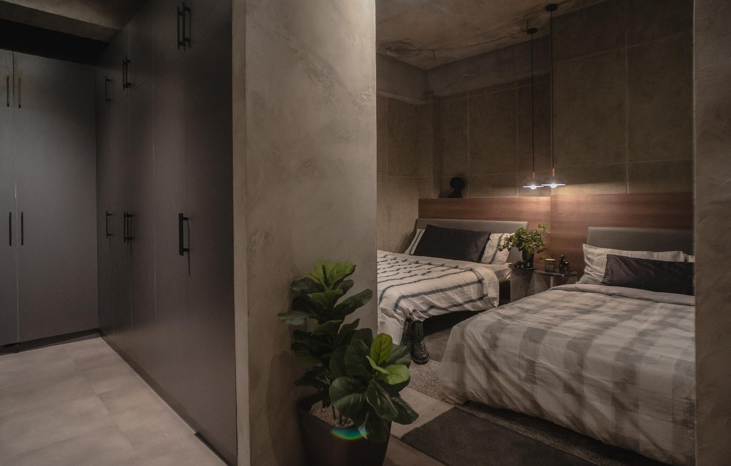 A dimly lit modern bedroom with two beds, a potted plant, and a row of wardrobes.